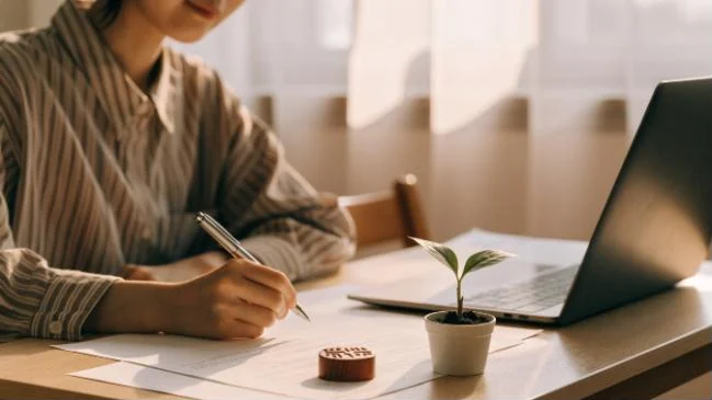 明るいデスクで書類に記入する人と、ノートパソコンと小さな芽の鉢